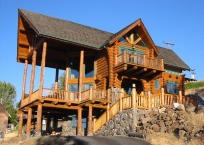 A two-story log cabin with a wraparound deck, stone foundation, and clear blue sky in the background.