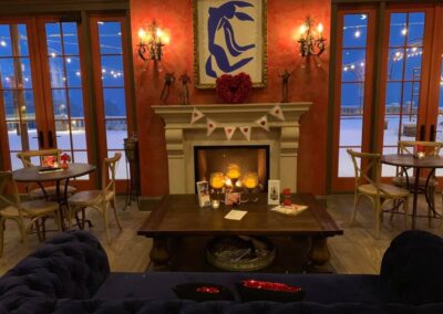 A cozy, warmly-lit room with a fireplace, candles, and valentine's day decorations, overlooking a snowy scene outside through large windows.