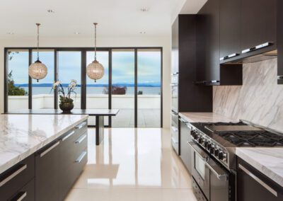 Modern kitchen with marble countertops, dark cabinets, and pendant lighting, featuring a scenic view through large windows.
