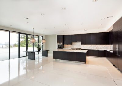 A modern kitchen with sleek dark cabinetry and island, equipped with stainless steel appliances and pendant lighting, featuring large windows with a view.
