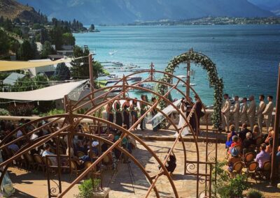 Outdoor wedding ceremony overlooking a scenic lake with guests seated under an ornamental archway.
