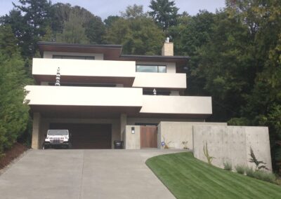 Modern multi-level house with a large garage and concrete driveway, surrounded by greenery.