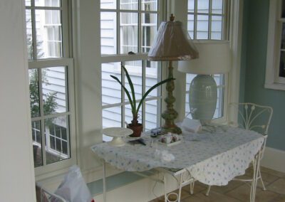 A small, cozy sunroom with a white metal table and chairs, a lamp, a potted plant, and a window view of the neighboring house.