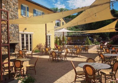 Outdoor café patio with umbrella shades, chairs, and tables set on a gravel surface, surrounded by a stucco building under a clear blue sky.