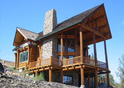 Log cabin with a large stone chimney and a wraparound wooden deck against a clear sky.